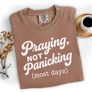 May include: A brown t-shirt with the words "Praying, Not Panicking (most days)" in white script. The shirt has a crew neck and a Comfort Colors tag. A cup of coffee on a wooden coaster and a small bouquet of dried flowers are also in the image.