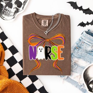 May include: Brown t-shirt with a Halloween-themed design. The word "NURSE" is spelled out with a ghost in place of the "U". The design includes a large orange and purple bow. A silver chain necklace is draped over the shirt.