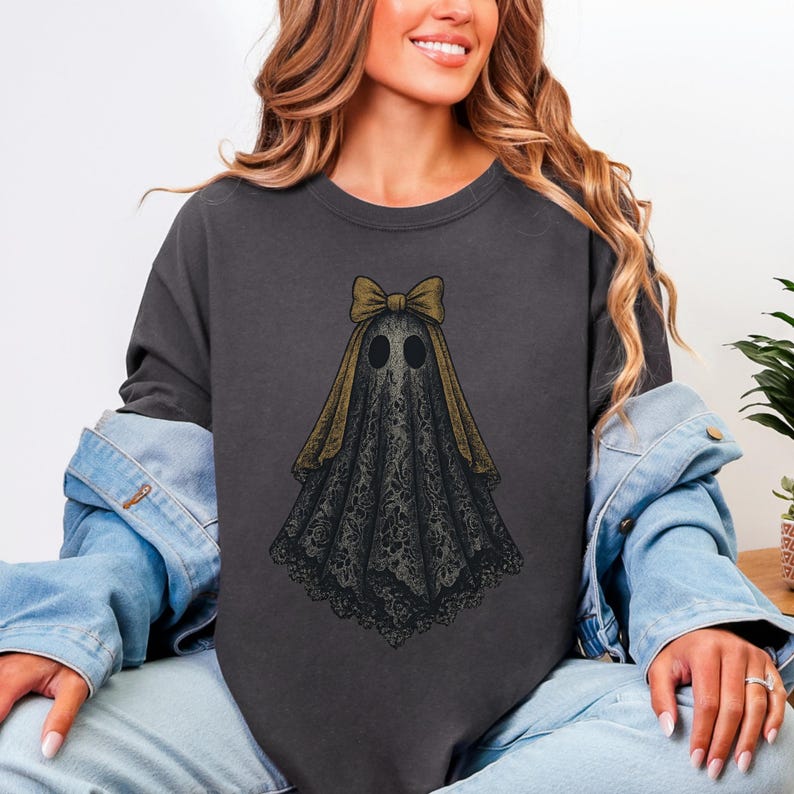 K&ouml;nnte beinhalten: Dunkelgraues Sweatshirt mit einem Geisterdesign. Der Geist ist mit einem schwarzen, spitzenartigen Leichentuch, einer goldenen Schleife und zwei schwarzen Augenl&ouml;chern dargestellt. Das Design ist auf der Vorderseite des Shirts zentriert und bietet eine gruselige, aber stilvolle &Auml;sthetik.