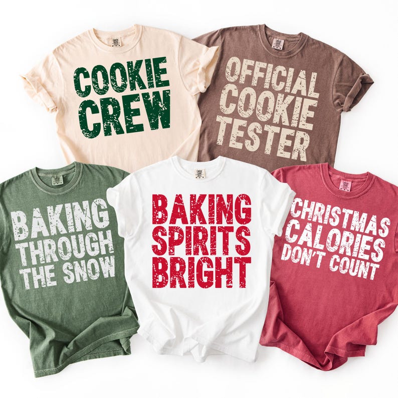 May include: Five short-sleeved t-shirts in various colors, each with a holiday-themed phrase in bold text. The shirts are beige, brown, white, green, and red. Phrases include "Cookie Crew", "Official Cookie Tester", and "Christmas Calories Don't Count".