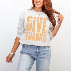 May include: A light gray sweatshirt with the words "GIVE THANKS" printed in a distressed orange font. The sweatshirt is long-sleeved and has a crew neck. The person is wearing blue jeans.