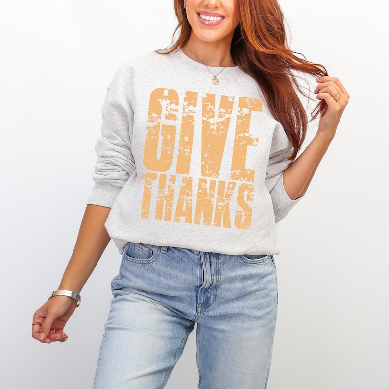 May include: A light gray sweatshirt with the words "GIVE THANKS" printed in a distressed orange font. The sweatshirt is long-sleeved and has a crew neck. The person is wearing blue jeans.