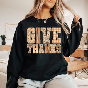 以下が含まれることがあります： 「GIVE THANKS」の文字がパッチワークデザインで施された黒いスウェットシャツ。文字はオレンジ、茶色、クリーム色の花柄、チェック柄、木目模様で埋められています。秋をテーマにした居心地の良いトップス。