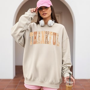 May include: A beige sweatshirt with the word "THANKFUL" in a decorative, patchwork style. The letters feature floral, plaid, and patterned designs in shades of brown, orange, and beige. The sweatshirt has long sleeves and a crew neck.