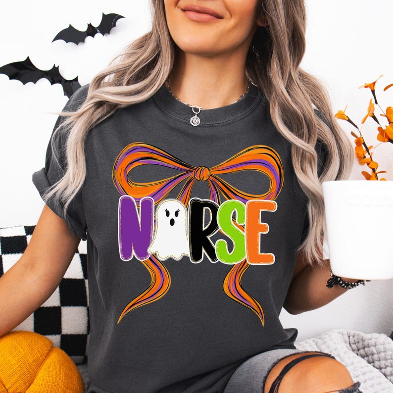 May include: Dark gray t-shirt with a Halloween-themed design. The design features the word "NURSE" in purple, white, green, and orange, with a ghost replacing the "O". An orange and purple bow is behind the text.