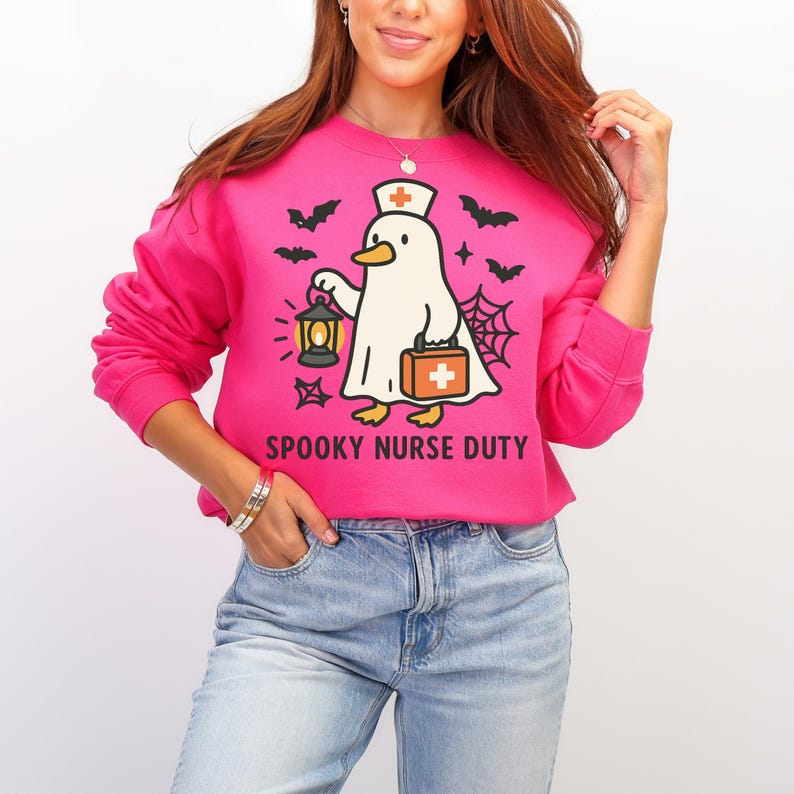 May include: A bright pink sweatshirt with the text "SPOOKY NURSE DUTY". The graphic features a ghost-like duck wearing a nurse's hat and carrying a lantern and medical bag, with bats and a spiderweb design.