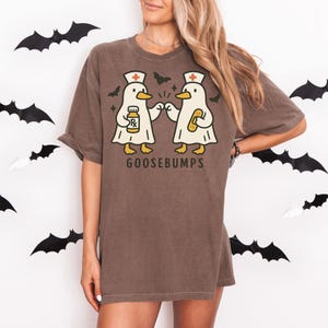 May include: Brown t-shirt with a graphic of two cartoon ducks wearing nurse hats and holding medical items. The text "GOOSEBUMPS" is printed below the ducks. Black bat cutouts decorate the background.