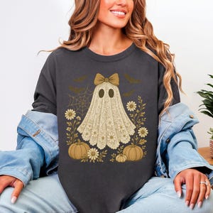 Puede incluir: Sudadera gris oscuro con un dise&ntilde;o de fantasma, lazo, flores, calabazas, murci&eacute;lagos y una telara&ntilde;a. El fantasma lleva un atuendo de encaje. Una camiseta gr&aacute;fica con tem&aacute;tica de Halloween.