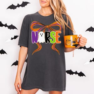 May include: Dark gray t-shirt with the word "NURSE" in colorful letters, a ghost, and an orange bow design. A Halloween-themed orange mug with a pumpkin and cat is held in the hand. Black bat decorations are in the background.