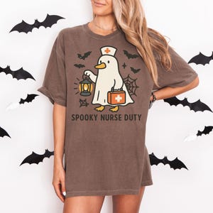 May include: Brown t-shirt with a Halloween-themed graphic. The design features a ghost wearing a nurse's hat and holding a lantern and medical bag. The text "SPOOKY NURSE DUTY" is printed below the graphic. Black bat cutouts decorate the background.
