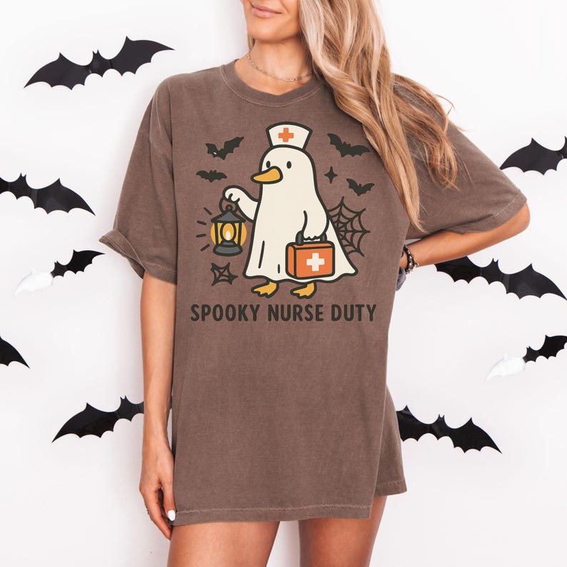 May include: Brown t-shirt with a Halloween-themed graphic. The design features a ghost wearing a nurse's hat and holding a lantern and medical bag. The text "SPOOKY NURSE DUTY" is printed below the graphic. Black bat cutouts decorate the background.