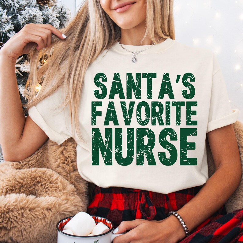 May include: Cream-colored t-shirt with the text "SANTA'S FAVORITE NURSE" in distressed green lettering. The person is wearing a silver necklace and a black bracelet. The person is also wearing red and black plaid pajama pants.