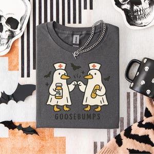 May include: Dark gray t-shirt with a graphic of two cartoon geese dressed as nurses, holding a prescription bottle and a bandage. The word "GOOSEBUMPS" is printed below the geese. A silver chain necklace is draped across the shirt.