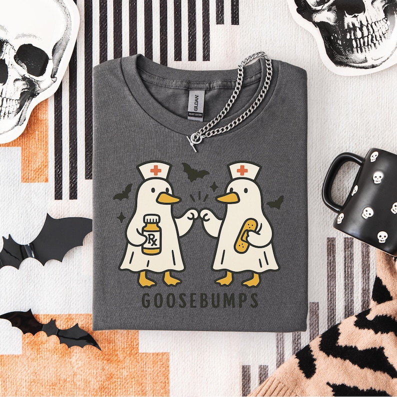 May include: Dark gray t-shirt with a graphic of two cartoon geese dressed as nurses, holding a prescription bottle and a bandage. The word "GOOSEBUMPS" is printed below the geese. A silver chain necklace is draped across the shirt.