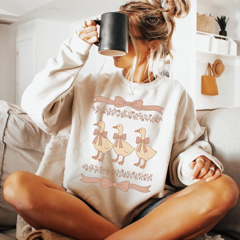 May include: A cream-colored sweatshirt with a graphic of three yellow ducks wearing pink bows. The ducks are lined up in a row and are surrounded by floral designs and pink ribbons.