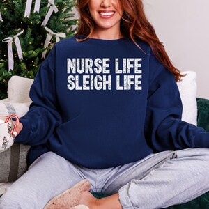 May include: Navy blue sweatshirt with the text "NURSE LIFE SLEIGH LIFE" in white block letters. The sweatshirt is a crew neck style and is made of a soft material. The image is of a person wearing the sweatshirt.