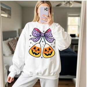 May include: White sweatshirt featuring a Halloween-themed graphic. The design includes two candy corn pieces with jack-o'-lantern faces, connected by a purple bow with polka dots. The design is accented with small star shapes.