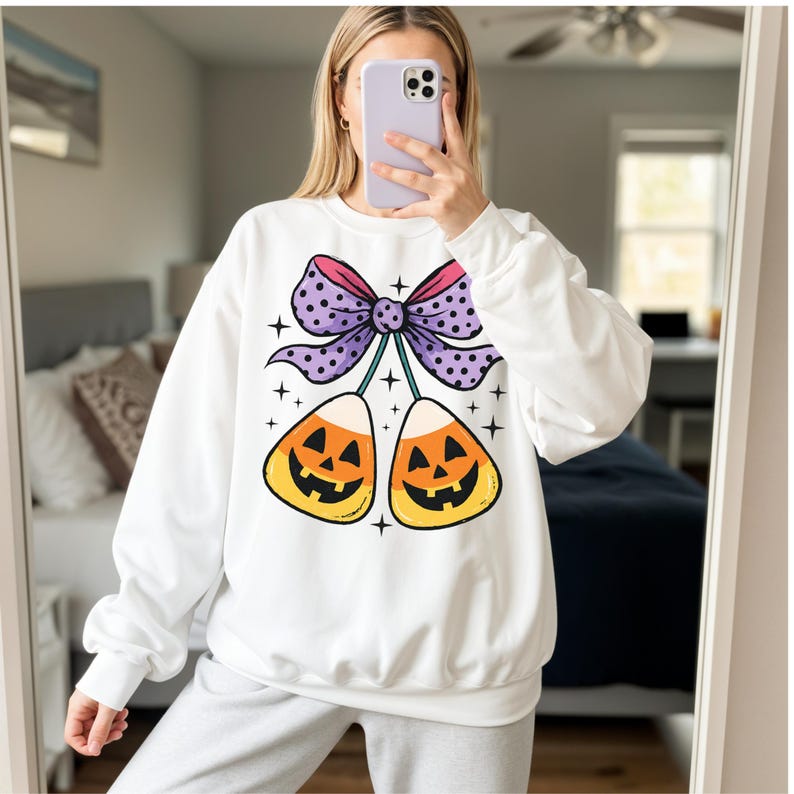 May include: White sweatshirt featuring a Halloween-themed graphic. The design includes two candy corn pieces with jack-o'-lantern faces, connected by a purple bow with polka dots. The design is accented with small star shapes.