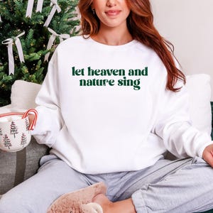 Puede incluir: Sudadera blanca de cuello redondo con el texto "let heaven and nature sing" en verde. La sudadera se usa con pantalones de ch&aacute;ndal grises. Un &aacute;rbol de Navidad est&aacute; en el fondo, y una taza con un dise&ntilde;o de &aacute;rbol de Navidad se sostiene.