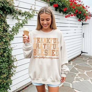 May include: A beige sweatshirt with the words "THANKFUL GRATEFUL BLESSED" in a patchwork design. The text "God is faithful in every season" is below. The person is holding an ice cream cone.