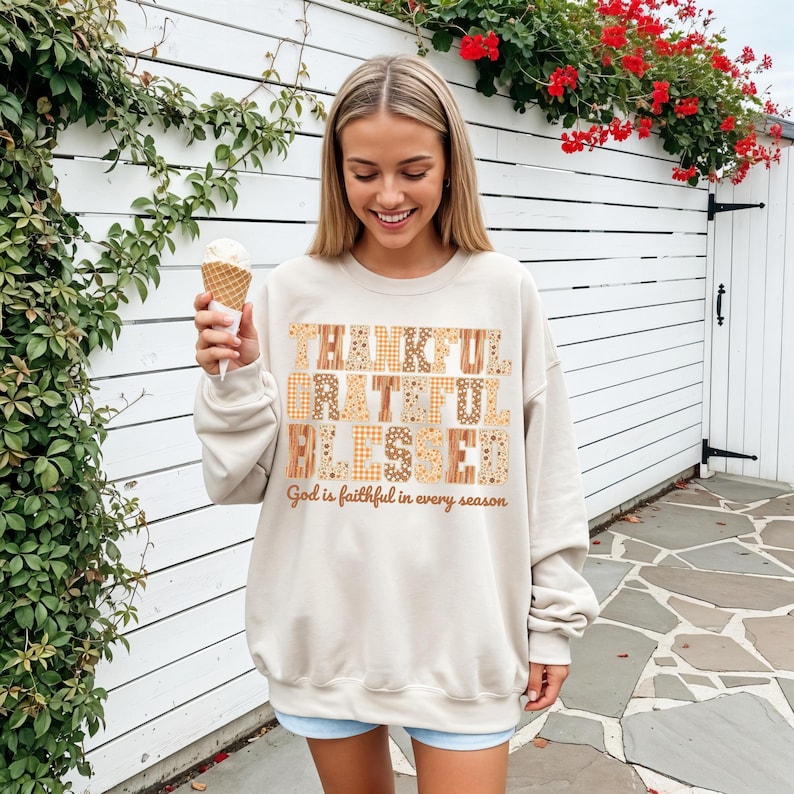 May include: A beige sweatshirt with the words "THANKFUL GRATEFUL BLESSED" in a patchwork design. The text "God is faithful in every season" is below. The person is holding an ice cream cone.