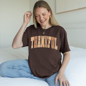 May include: A brown t-shirt with the word "THANKFUL" in a patchwork design across the front. The letters are in shades of orange, brown, and cream. The shirt is short-sleeved and worn by a person.