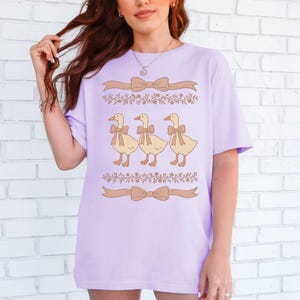May include: A lavender t-shirt with a graphic of three yellow ducks wearing brown bows. The ducks are lined up in a row and are surrounded by brown floral vines and bows.