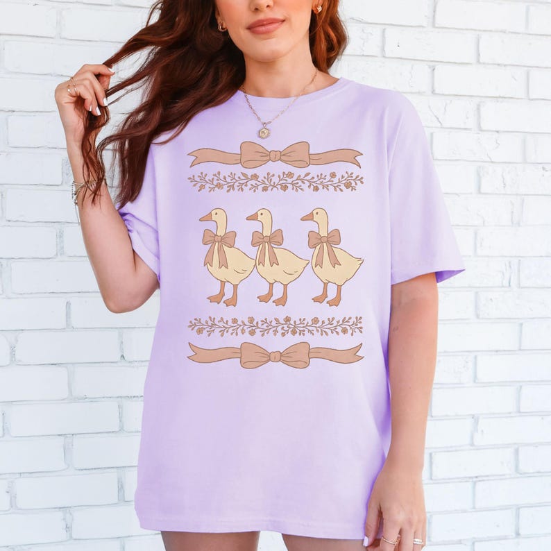 May include: A lavender t-shirt with a graphic of three yellow ducks wearing brown bows. The ducks are lined up in a row and are surrounded by brown floral vines and bows.