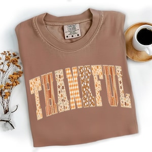 May include: A brown Comfort Colors t-shirt with the word "THANKFUL" in a patchwork design. The letters feature floral, plaid, and wood-grain patterns. A cup of coffee and dried flowers are in the background.