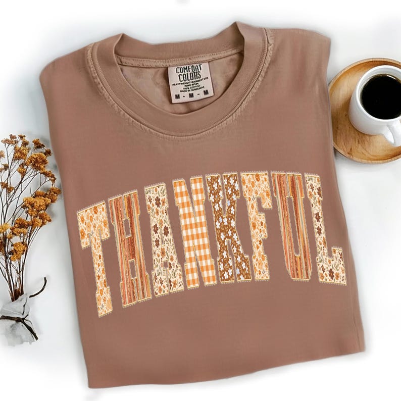 May include: A brown Comfort Colors t-shirt with the word "THANKFUL" in a patchwork design. The letters feature floral, plaid, and wood-grain patterns. A cup of coffee and dried flowers are in the background.