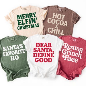 May include: Five short-sleeved t-shirts in various colors, each with a holiday-themed phrase in bold lettering. Phrases include "Merry Elfin' Christmas," "Hot Cocoa & Chill," and "Santa's Favorite Ho."