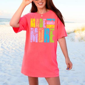 May include: A coral-colored t-shirt with the words "MADE MORE" in a colorful, distressed font. The text is in yellow, blue, green, purple, and orange. The shirt is worn on a beach.