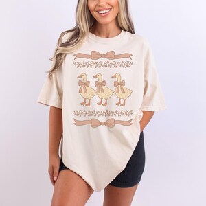 May include: A cream colored t-shirt with a graphic of three ducks wearing bows. The ducks are in a line and are facing the right. The graphic is surrounded by floral designs and ribbons.