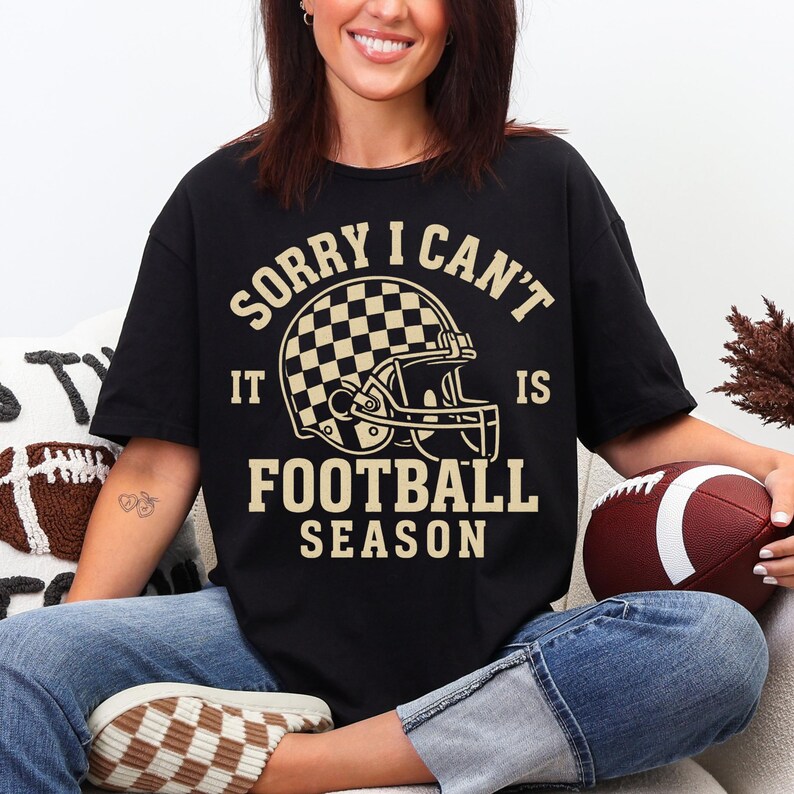 K&ouml;nnte beinhalten: Schwarzes T-Shirt mit dem Text "SORRY I CAN'T IT IS FOOTBALL SEASON" und einer Footballhelm-Grafik. Der Helm hat ein Schachbrettmuster. Ein brauner und wei&szlig;er Football ist ebenfalls zu sehen.