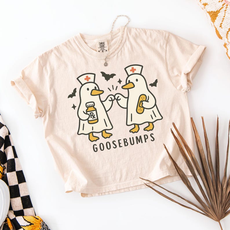 May include: Cream-colored t-shirt featuring a graphic of two cartoon geese dressed as nurses, with the text "GOOSEBUMPS" below. One goose holds a prescription bottle, the other a bandage. Bats and stars are also in the design. A silver necklace is visible.