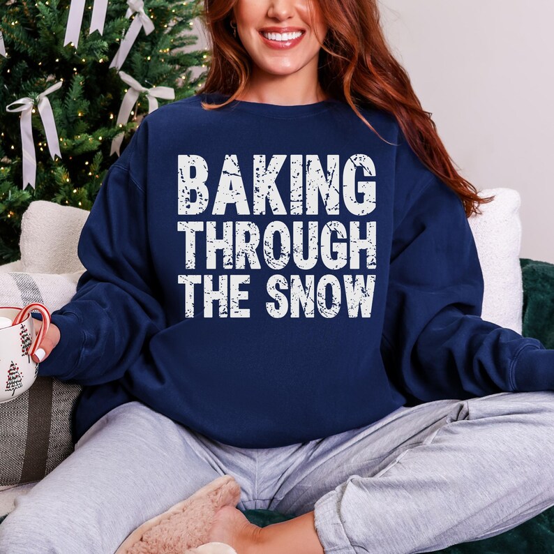May include: Navy blue sweatshirt with the text "BAKING THROUGH THE SNOW" in white. The sweatshirt is a crew neck style. The image shows a person wearing the sweatshirt, seated in front of a Christmas tree.
