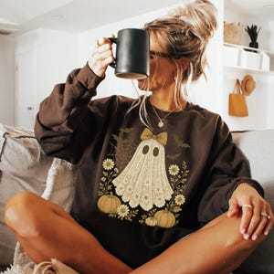 Puede incluir: Una sudadera marr&oacute;n con tem&aacute;tica de Halloween con un dise&ntilde;o de fantasma con un lazo, rodeado de calabazas, flores y murci&eacute;lagos. El fantasma lleva un lazo. La persona sostiene una taza negra.