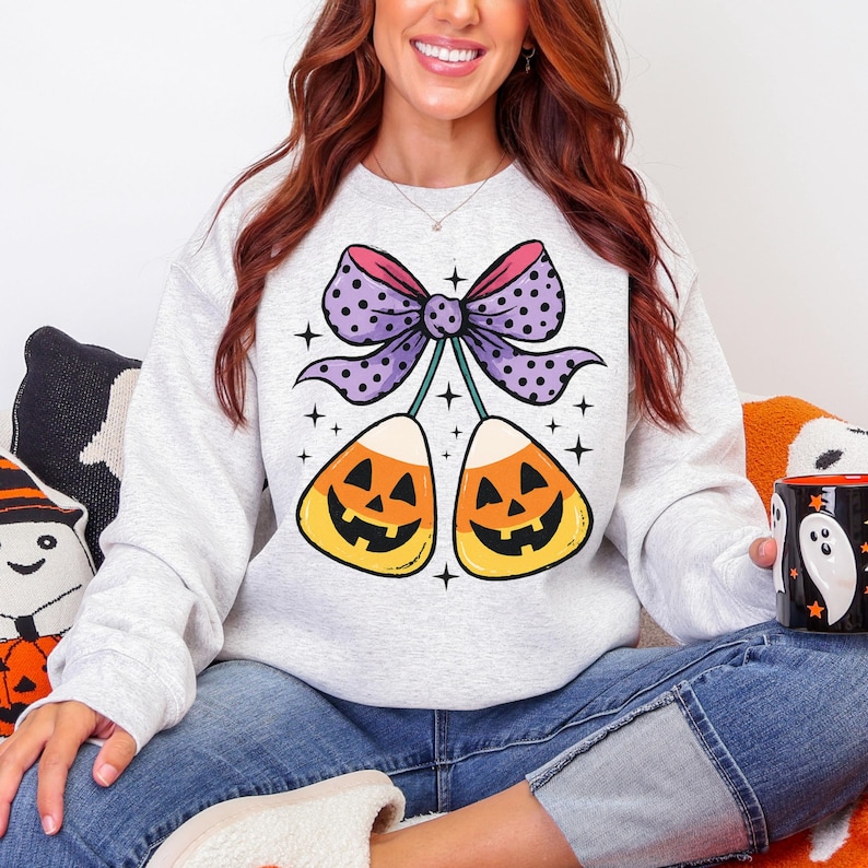 May include: A light gray sweatshirt featuring a Halloween-themed graphic. The design includes two candy corn with jack-o'-lantern faces, connected by a purple bow with polka dots. The sweatshirt is a comfortable, casual style.