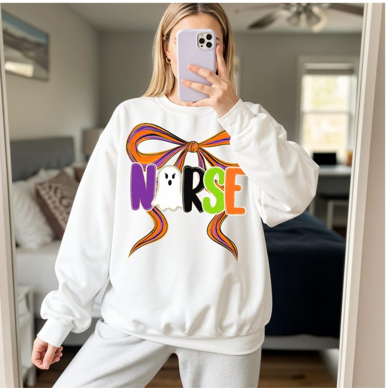 May include: White sweatshirt with a Halloween-themed design. The word "NURSE" is spelled out in purple, black, green, and orange letters, with a ghost graphic in the center. An orange and purple bow design is above the word.