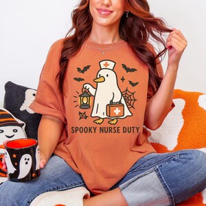 May include: Rust-colored t-shirt with a Halloween-themed graphic. The design features a cartoon duck wearing a nurse's cap and carrying a lantern and medical bag, with bats and a spiderweb. The text "SPOOKY NURSE DUTY" is below the graphic.