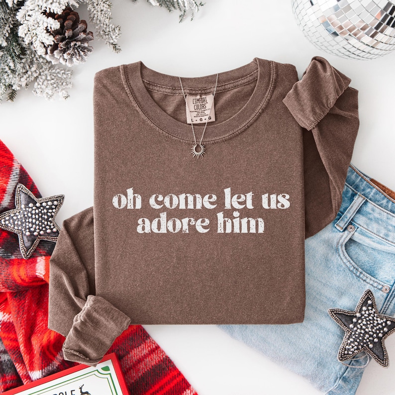 Puede incluir: Una camiseta marr&oacute;n de manga larga con la frase blanca "oh come let us adore him". Se ve un collar plateado con un colgante de sol. La camiseta est&aacute; doblada con unos vaqueros azules y adornos navide&ntilde;os.