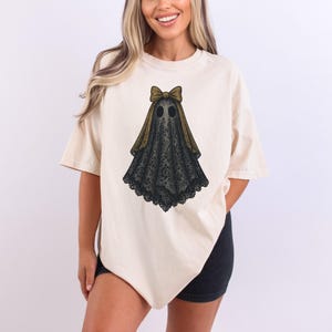 K&ouml;nnte beinhalten: Cremefarbenes T-Shirt mit Geisterdesign. Der Geist ist mit einem schwarzen, spitzenartigen K&ouml;rper, einer goldenen Schleife und zwei schwarzen Augenl&ouml;chern dargestellt. Das Shirt hat kurze &Auml;rmel und eine lockere Passform.