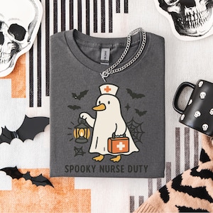 May include: Dark gray t-shirt with a cartoon ghost penguin in a nurse's hat, holding a lantern and medical bag. The text "SPOOKY NURSE DUTY" is below the graphic. Halloween-themed shirt with bats and a spiderweb design.
