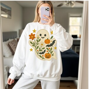 May include: White sweatshirt with a smiling ghost design, orange pumpkins, bats, flowers, and stars. The ghost is surrounded by floral elements. The design is embroidered in orange, green, and yellow.