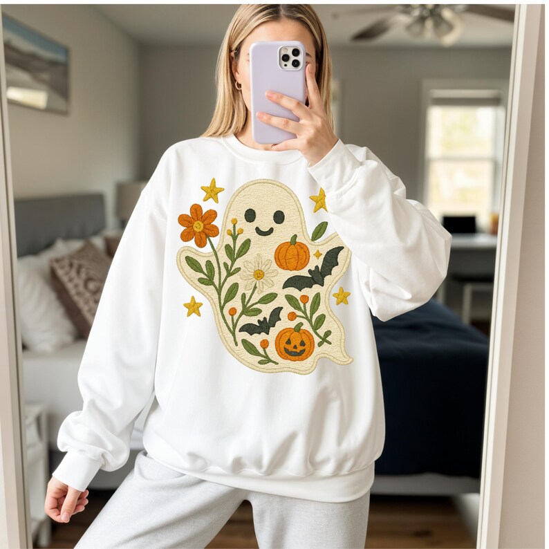May include: White sweatshirt with a smiling ghost design, orange pumpkins, bats, flowers, and stars. The ghost is surrounded by floral elements. The design is embroidered in orange, green, and yellow.