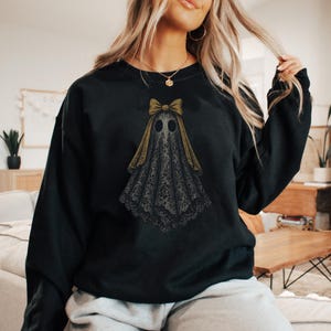 K&ouml;nnte beinhalten: Schwarzes Sweatshirt mit Geisterdesign. Der Geist hat eine goldene Schleife und ein schwarzes spitzenartiges Gewand. Das Design hat eine Vintage-, Gothic-&Auml;sthetik, ideal f&uuml;r Halloween oder den Alltag.