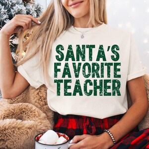 May include: Cream-colored t-shirt with the text "SANTA'S FAVORITE TEACHER" in green block letters. The shirt is worn with red and black plaid pajama pants. A mug with marshmallows is visible.