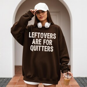Pu&ograve; includere: Una felpa marrone scuro con la scritta bianca "LEFTOVERS ARE FOR QUITTERS". La felpa &egrave; indossata con un berretto da baseball bianco e delle cuffie. La persona tiene in mano un bicchiere di plastica trasparente con una cannuccia.