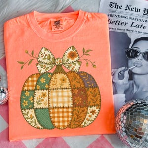 Könnte beinhalten: Korallfarbenes T-Shirt mit einem Patchwork-Kürbis-Design und einer großen Schleife. Der Kürbis besteht aus verschiedenen gemusterten Stoffquadraten in Orange, Grün und Gelb. Das Shirt hat einen Rundhalsausschnitt und kurze Ärmel, ideal für den Herbst.
