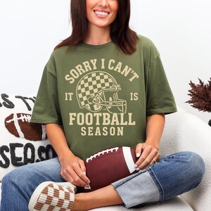K&ouml;nnte beinhalten: Olivgr&uuml;nes T-Shirt mit dem Text "SORRY I CAN'T IT IS FOOTBALL SEASON" und einer Footballhelm-Grafik. Die Person h&auml;lt einen braunen und wei&szlig;en Football. Das Shirt wird mit Jeans und braunen und wei&szlig;en Schuhen kombiniert.